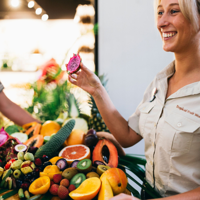 A Tropical Fruit World guide holds a slice of dragon fruit beside a colourful display of tropical fruit